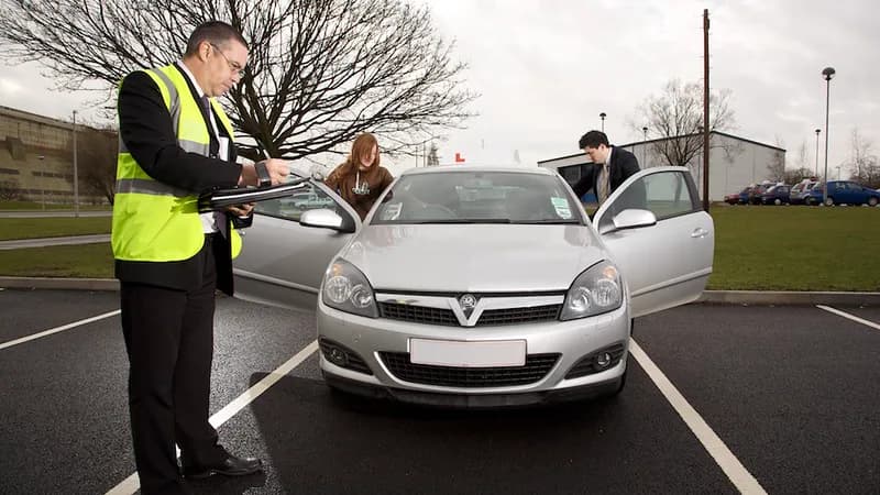 A driving examiner with a clipboard beside a learner car as the learner and instructor get in at the start of a practical driving test